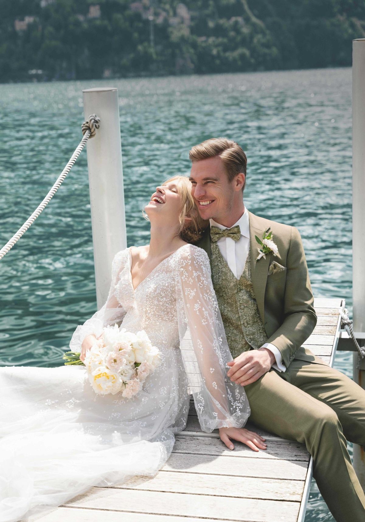 Ein Hochzeitspaar, das auf einem Steg an einem See sitzt, wobei die Braut einen Blumenstrauß hält.