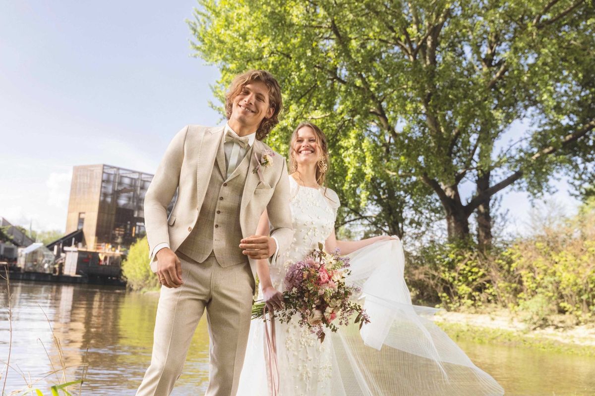 Braut und Bräutigam halten sich an einem Fluss mit Bäumen und einem Gebäude im Hintergrund an den Händen.